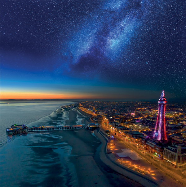 Aerial view of seafront and tower illuminated at night with a starry sky.
