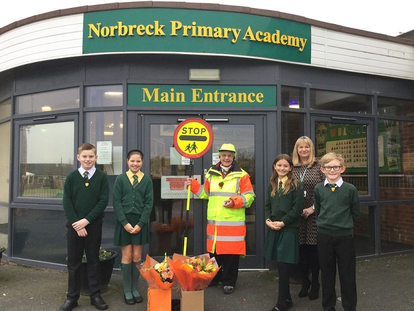 School crossing patrol officer with pupils and a teacher, stood outside a school with flowers