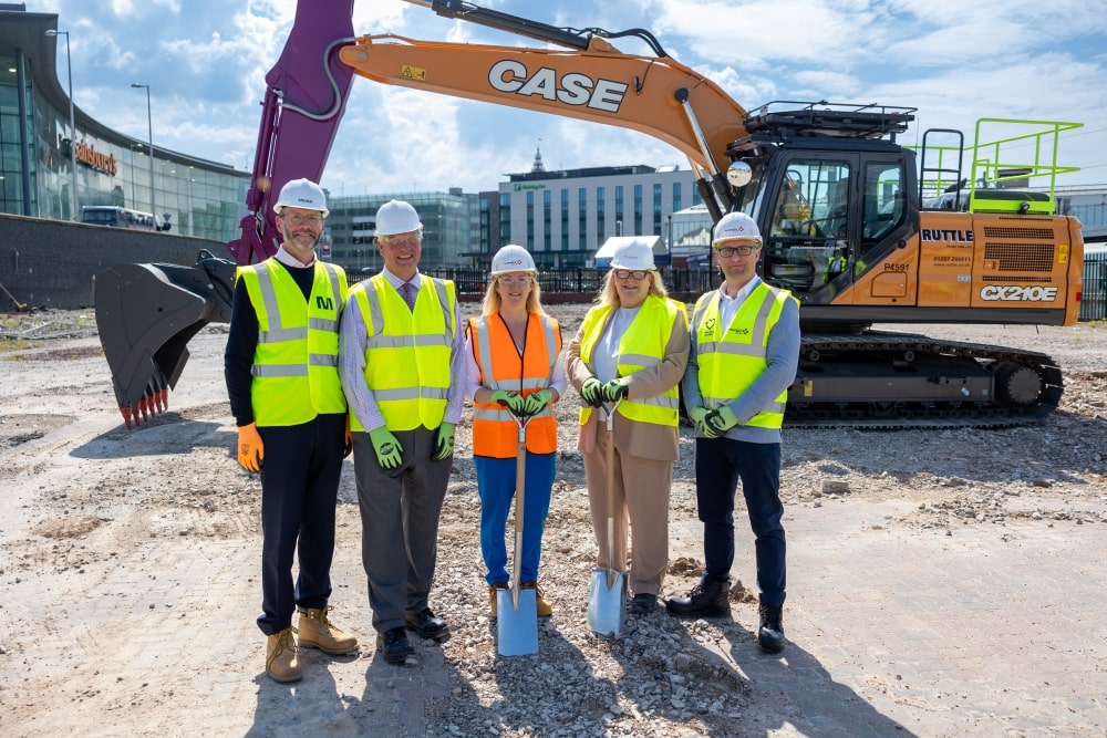 A photo of people stood on a construction site holding a spade. A digger, buildings and Blackpool Tower are in the background