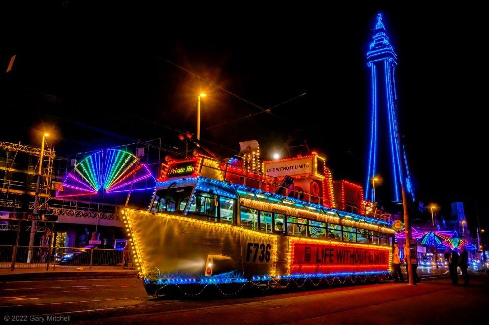 A photograph of an illuminated tram on Blackpool Promenade with Blackpool Tower in the background