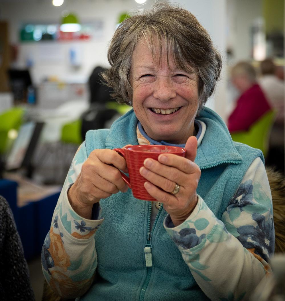 Lady sits down with a hot drink smiling at the camera at a library event.