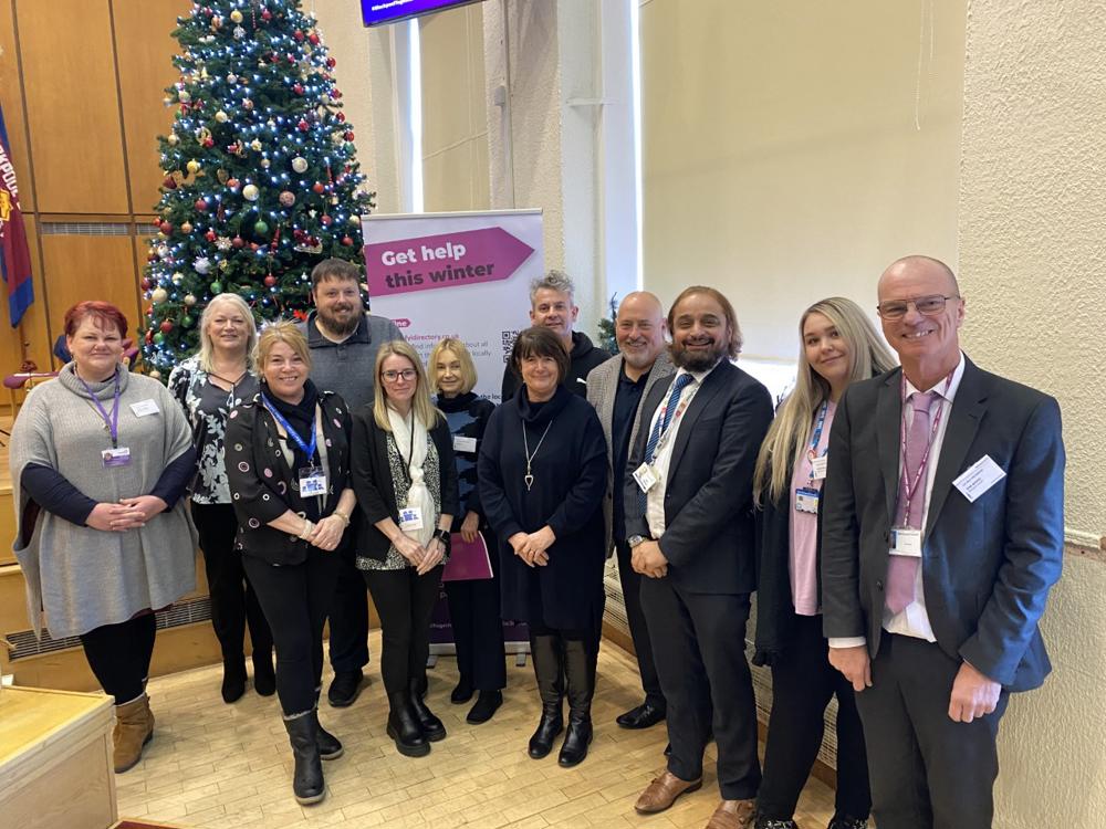 Photograph featuring representatives from council staff and community groups involved in the Blackpool Together Campaign. They stand in front of a Christmas tree and a branded banner, which reads "get help this winter". 