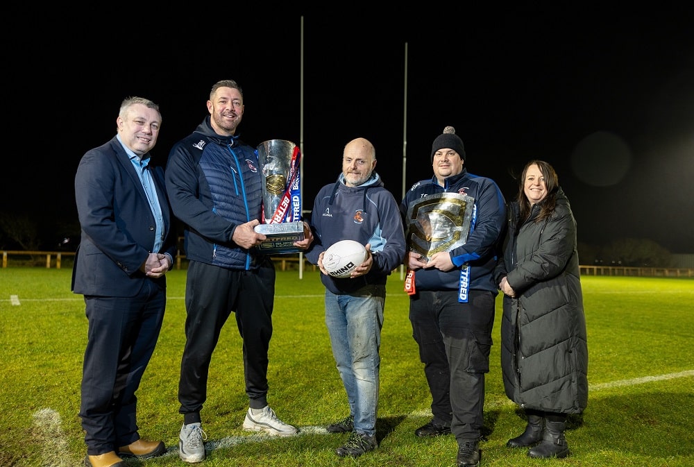 A group of five people with rugby balls and trophies on a grass pitch with rugby posts behind