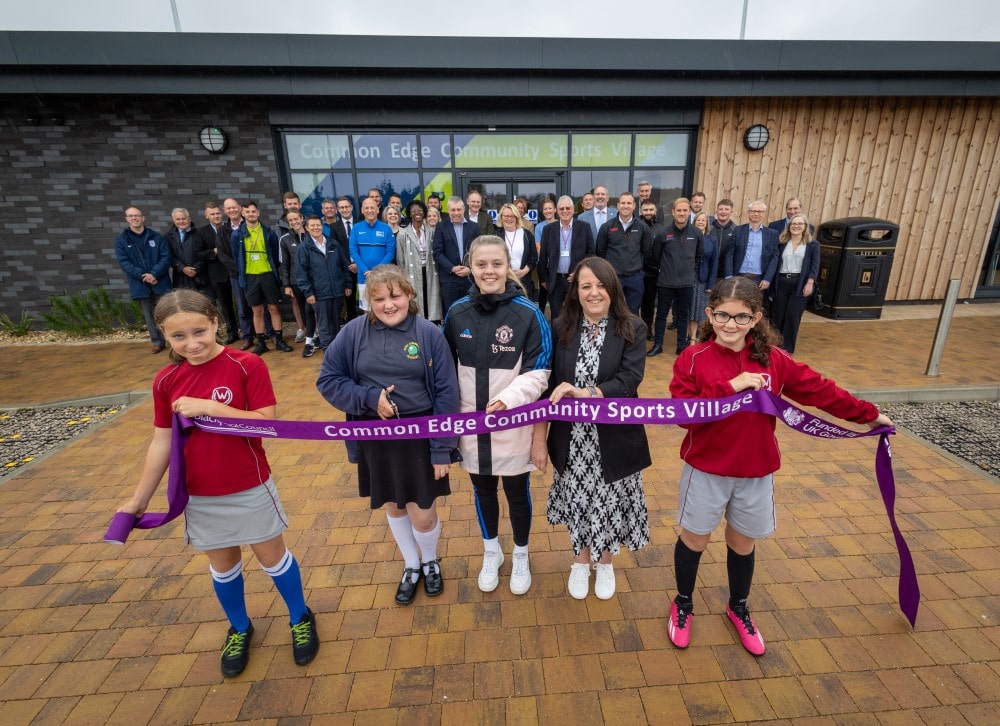 Five girls holding a ribbon saying Common Edge Community Sports Village with a group of people and a new building behind.