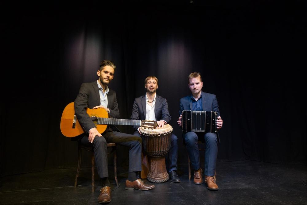 Three musicians sit on chairs, one holding a classical guitar, one a drum and the other an accordion. 