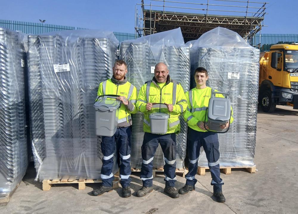Three people stood holding food waste caddies in front of stacks of packaged caddies.