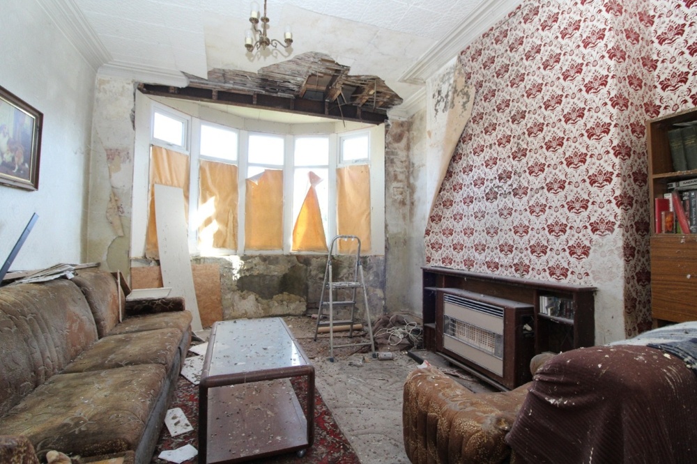 Image shows a damaged living room, with part of the ceiling having fallen down and pigeon droppings across the surfaces