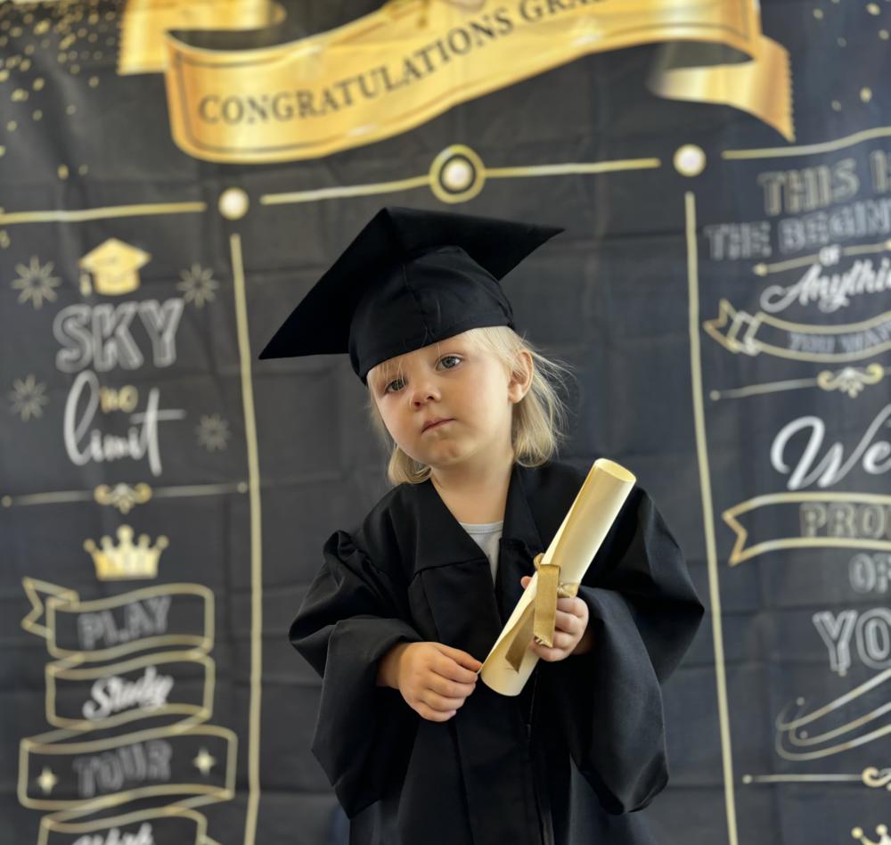 Two year old boy with blonde hair poses for picture with a graduation cap and gown with a black photo back drop