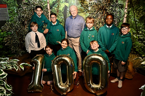 Group of school children stood with wax figure of Sir David Attenborough with balloons that make up the number 100.
