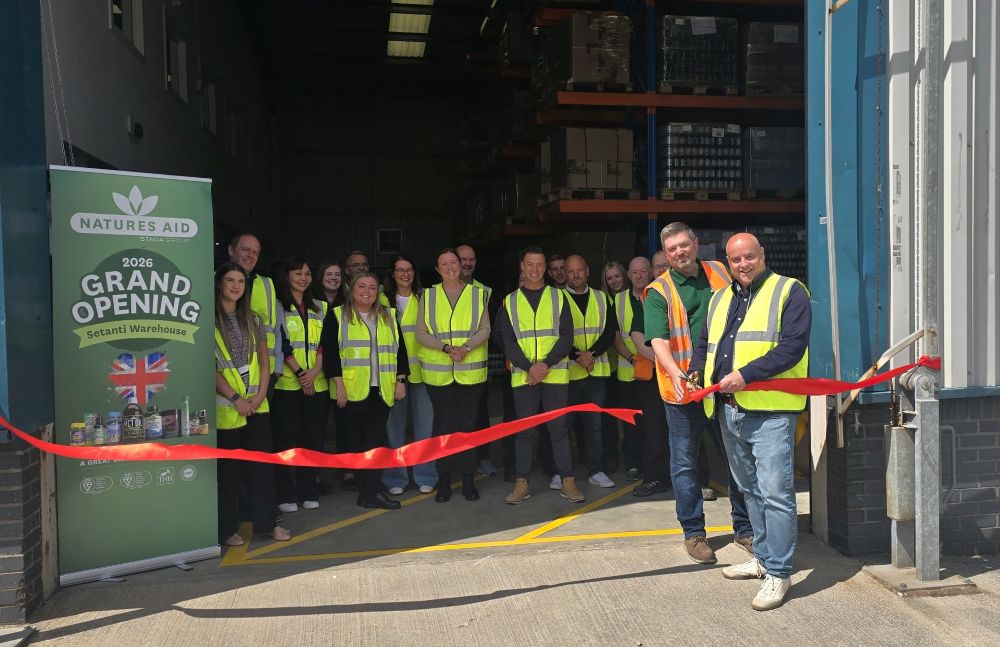 A group of workers gathered at the entrance to a warehouse wearing yellow high-visibility vests. Two workers are cutting a re ribbon with scissors to announce the opening of the Natures Aid warehouse.