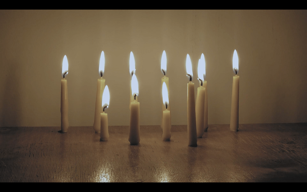 A slow and burning hope, (still from moving image work) by RA WALDEN.  11 candles sit burning on wooden surface.