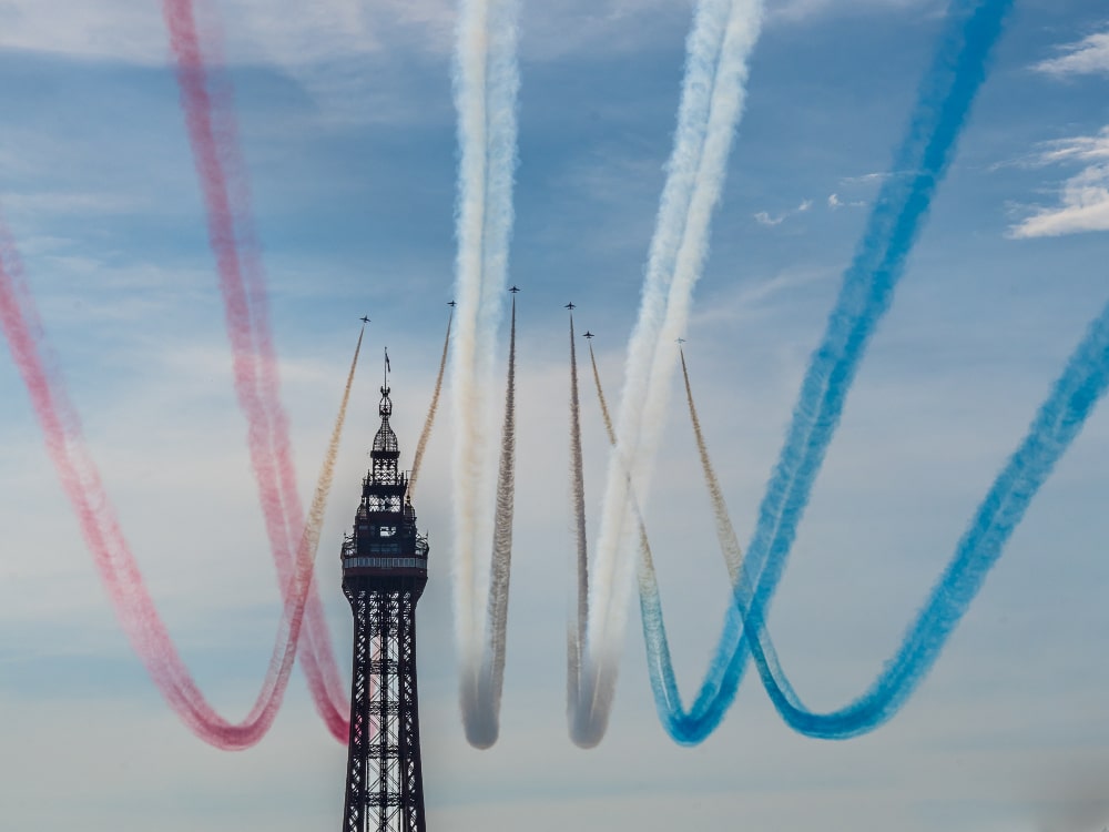 The RAF Red Arrows flying past Blackpool Tower with red, white and blue vapour trails coming out of the jets.