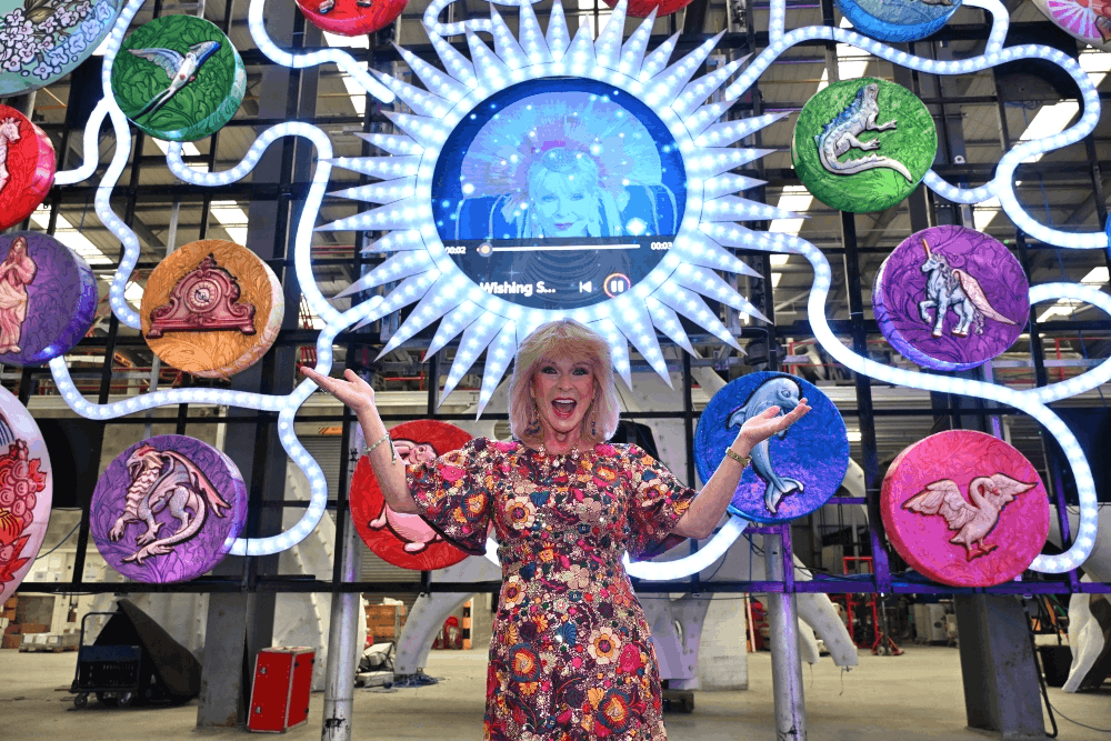Woman stood in front of light design with arms outstretched and open-mouth smile.
