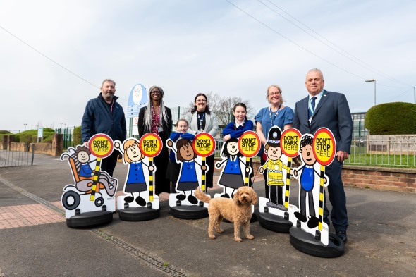 A group of people with a dog stand on a sidewalk next to cartoon cutouts displaying "Don't park here" signs.