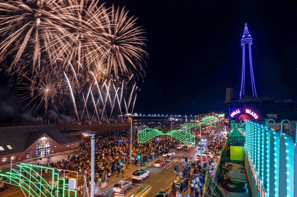 Large crowd standing on promenade watching fireworks display and illuminated tower and displays over roadway 