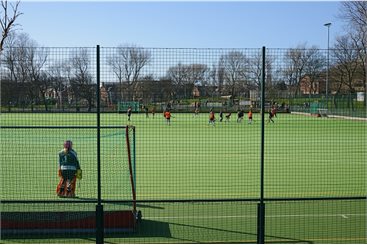 Sports pitches surrounded by fencing with a game of Hockey taking place on the pitch