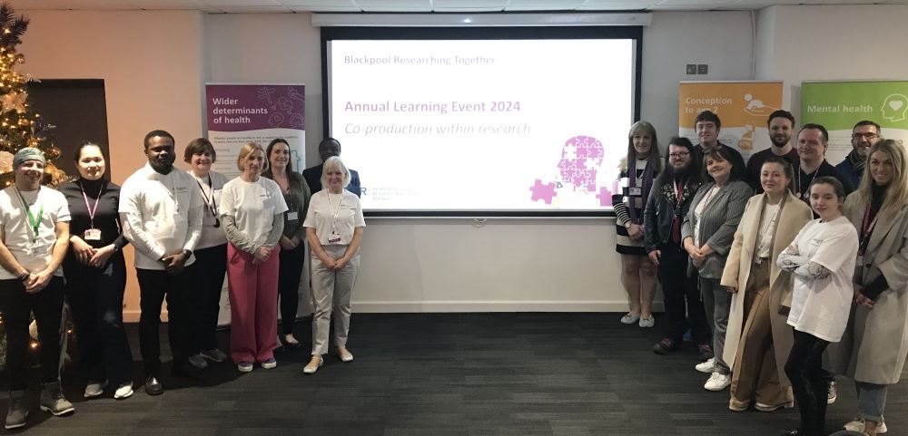 Blackpool Researching Together team surrounding their presentation slideshow screen at the learning event.