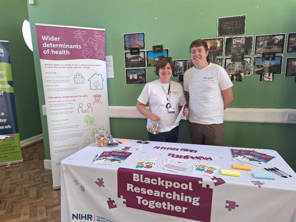 Man and woman stood behind a table containing merchandise at an event. They are wearing white t-shirts which feature purple Blackpool Researching Together branding.