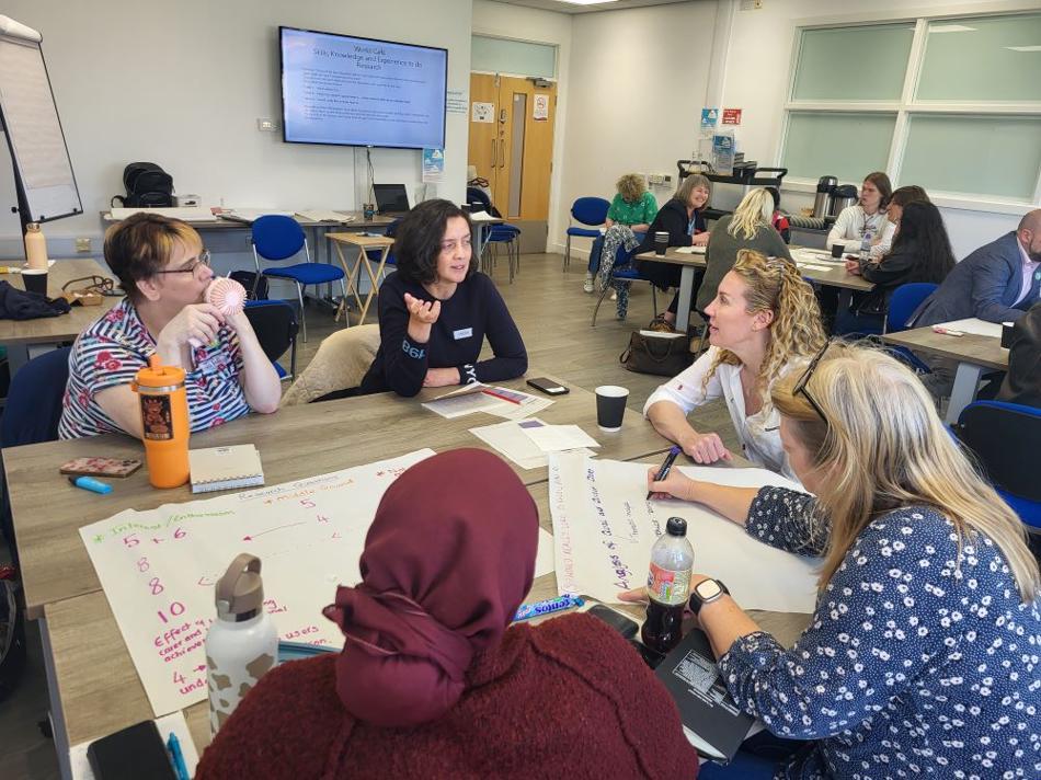 Groups of people sat around tables working collaboratively. The tables have poster paper and pens on.