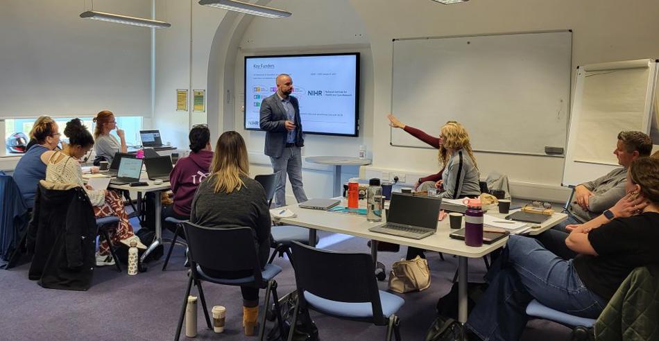 People grouped into tables watching a man present at the front of the room.