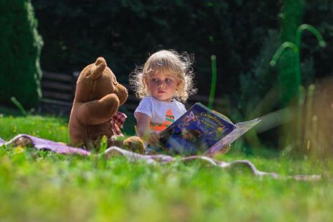 Girl toddler reading a book with her teddy bear on the grass in the park