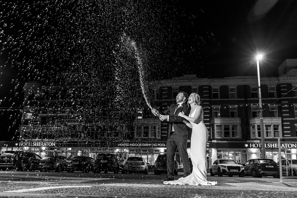 Bride and groom opening a bottle of champagne outside a hotel.