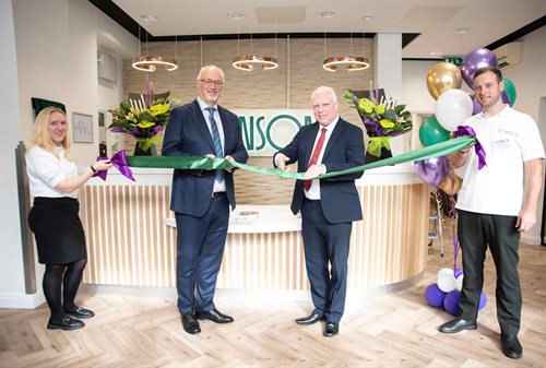 Four people smile at the camera whilst holding a green ribbon in front of a reception desk. One man holds a pair of scissors ready to cut the ribbon
