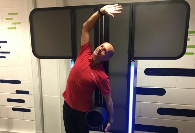 Instructor Phil using the Express fitness Flex machine with one hand up in the air the other on the machine