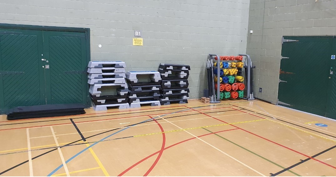 Exercise class equipment including mats, bodypump weights and step boxes stacked up to one side of the sports hall with socially distanced markings on the floor